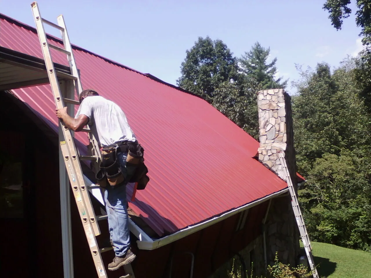 Roofer on a ladder beside a red metal roof and stone chimney — Soffit & Fascia Repair by Roofing Brothers in Westley